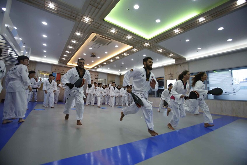A group of people in white martial arts uniforms practicing in a well-lit indoor facility with blue mats. 4