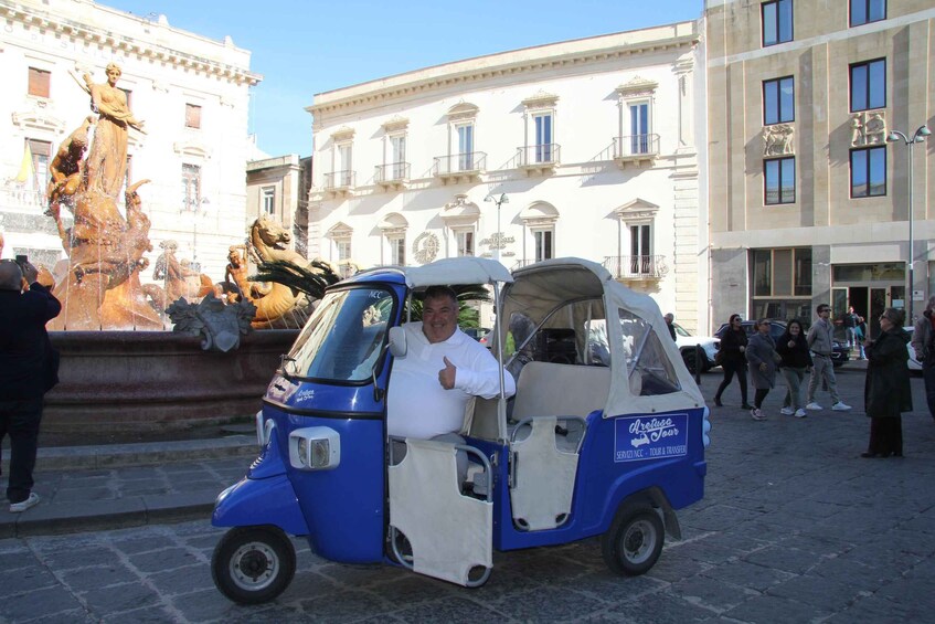 A blue and white tuk-tuk parked in front of a historic building with a fountain. 1