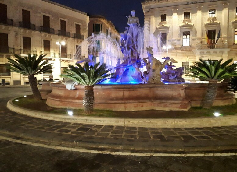 A nighttime fountain with water jets and sculptures, surrounded by palm trees and classical architecture. 3