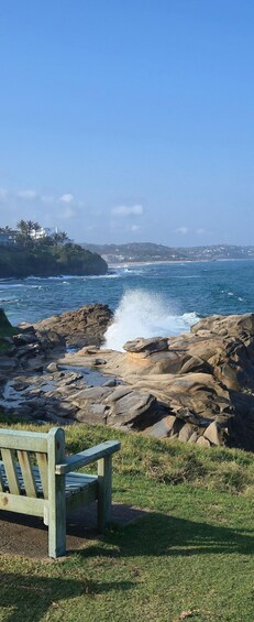 A rocky shoreline with waves crashing, a bench, and a clear blue sky. 3