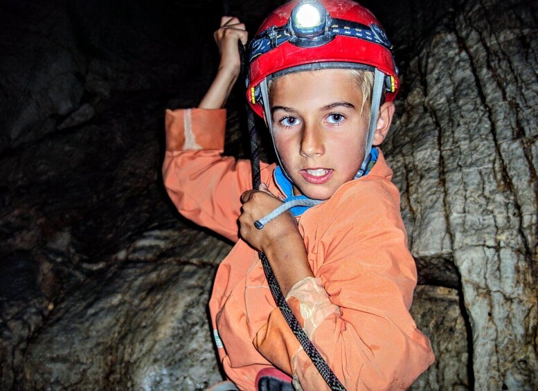A person in an orange jacket and red helmet with a headlamp, sitting in a cave. 1