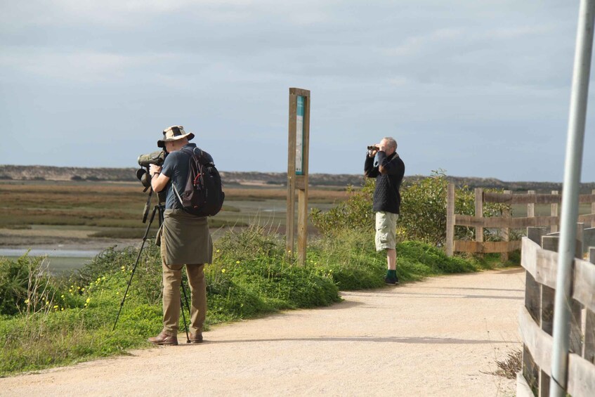 Two people on a dirt path with a camera and binoculars, a wooden signpost, and a fence. 1