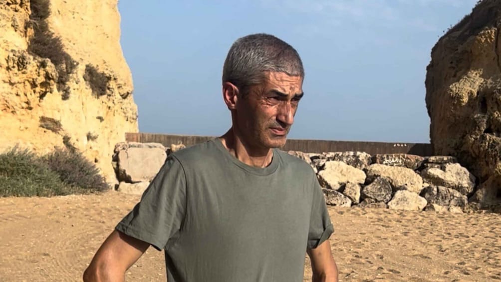 A man standing outdoors with a rocky cliff and a stone wall in the background. 3