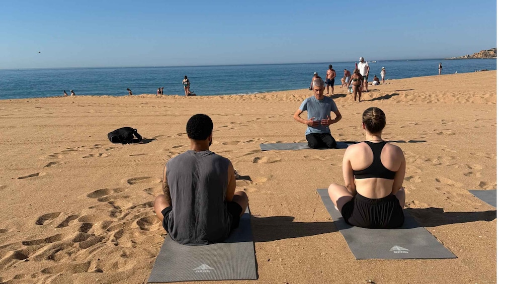 People sitting on yoga mats on a sandy beach with the ocean in the background. 1