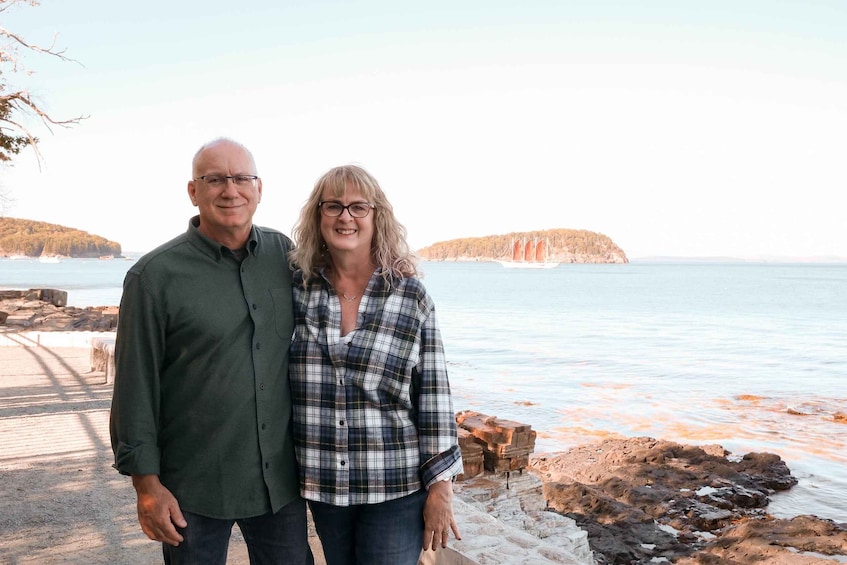Two people standing by a rocky shoreline with a body of water and distant landforms in the background. 1