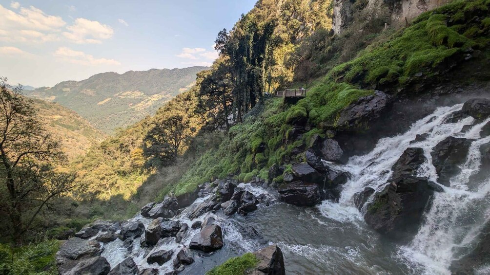 A waterfall cascading over rocks, surrounded by lush greenery and a forested valley. 2