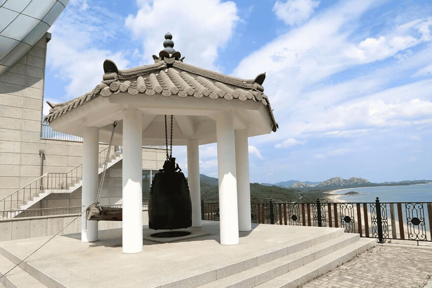 A traditional pavilion with a hanging bell, a staircase, and a view of a beach and mountains. 4