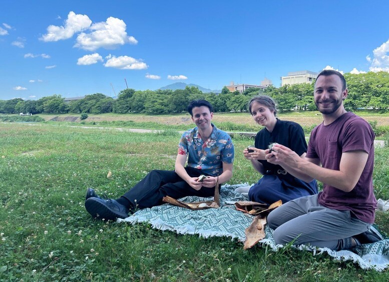Three people sitting on a blanket in a grassy field with trees and buildings in the background. 4
