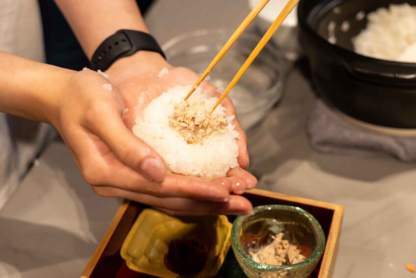 A person holding a bowl of rice with chopsticks, with a container of sauce and a bowl of rice in the background. 5