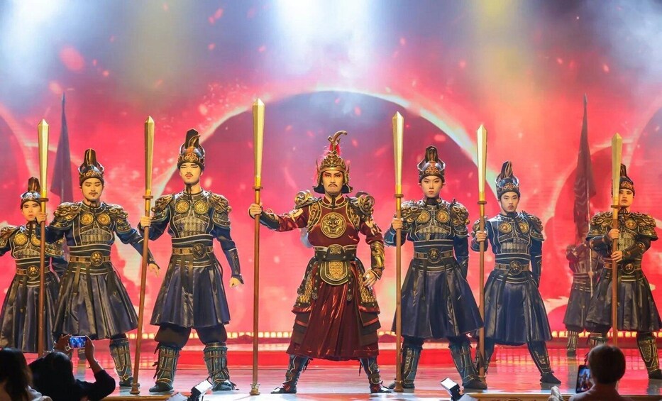 Eine Gruppe von Darstellern in traditioneller Tracht und Rüstung, mit Schwertern, vor einem roten Bühnenhintergrund. 3