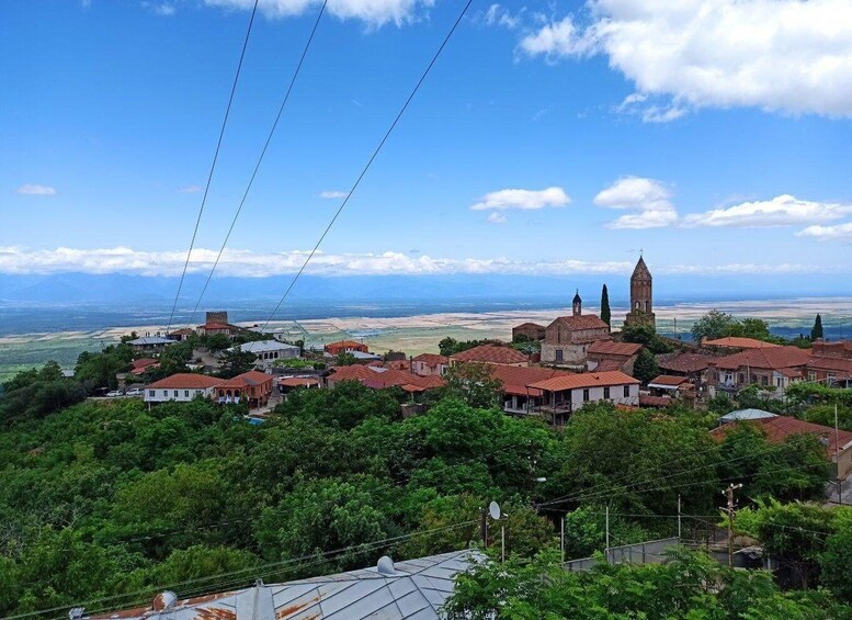 A coastal town with red-roofed buildings, a prominent church tower, and a view of the sea and distant mountains. 1