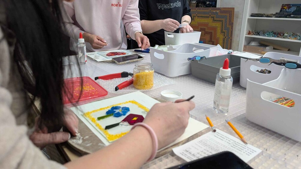 A person working on a craft project with various materials like beads, a jar of yellow beads, and a red tool on a table. 1