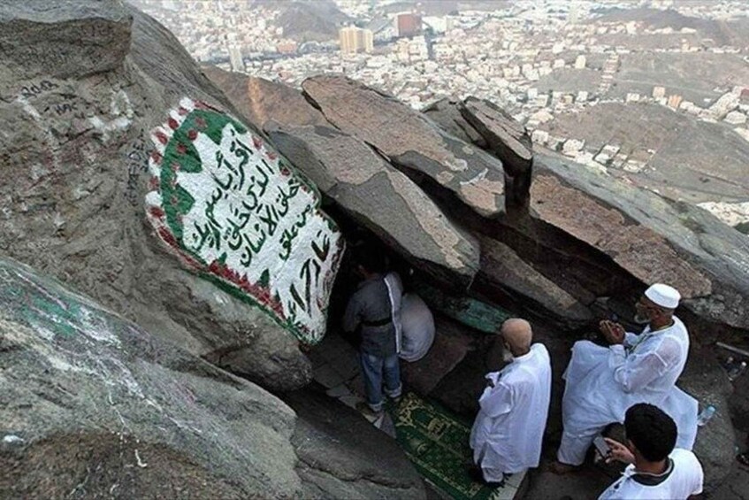 A rocky terrain with a carved inscription, a group of people, and a cityscape in the background. 2