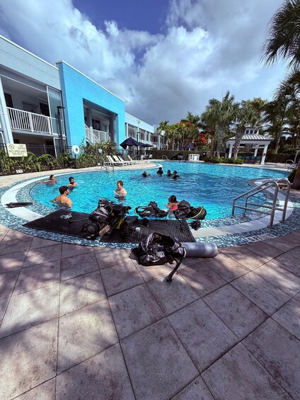 A swimming pool with people in the water, a diving board, and a building in the background. 1