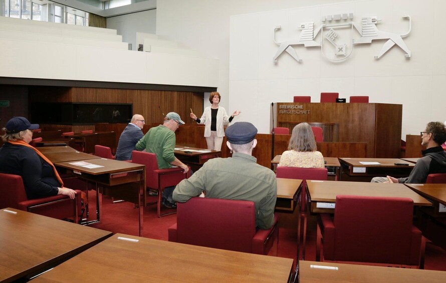 A lecture hall with attendees seated at tables, a lecturer at the front, and a wall with a white emblem. 4