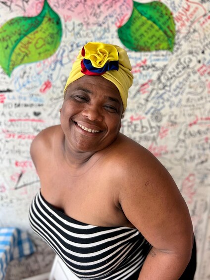 A person wearing a striped dress and a yellow headscarf with a blue and red flower, standing in front of a wall with colorful graffiti. 1