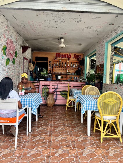 A restaurant with a sign reading 'Mama Rosa,' featuring a tiled floor, checkered tablecloths, wicker chairs, and a wall covered in handwritten notes. 2