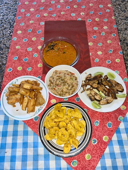 A table with a red patterned tablecloth, a bowl of soup, a plate of fried plantains, a plate of rice with peas, a plate of grilled fish, and a glass of sauce. 3