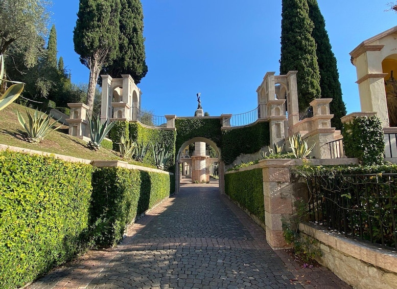 A cobblestone pathway flanked by neatly trimmed hedges and tall cypress trees, leading to a classical archway. 4