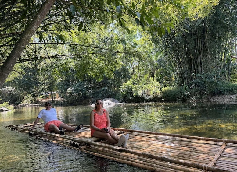 Two people sitting on a bamboo raft in a river surrounded by dense greenery. 2