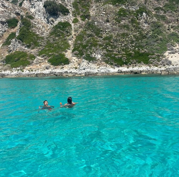 Two people swimming in clear turquoise water with a rocky coastline and greenery in the background. 4