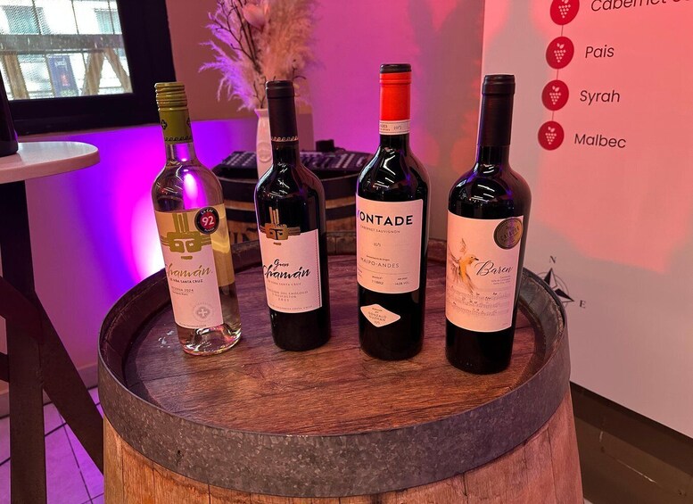 Four wine bottles on a wooden barrel with labels indicating Cabernet, Pais, Syrah, and Malbec varieties. 1
