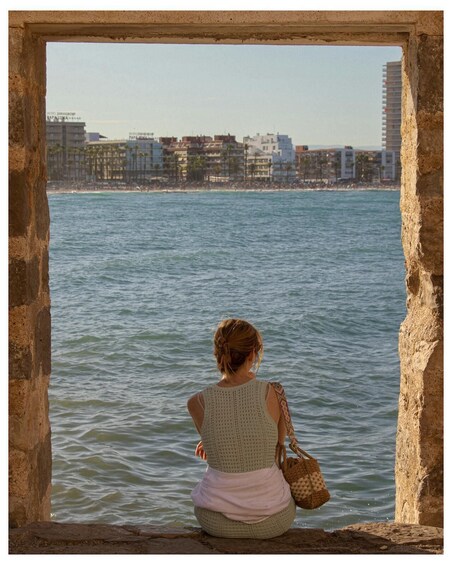 A person sitting by a body of water, overlooking a cityscape with buildings and a beach. 3