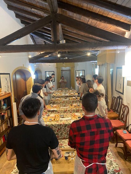 A group of people in a room with a long table set for a meal, wooden beams on the ceiling, and framed artwork on the walls. 2
