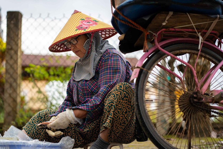 A person sitting on a stool, wearing a patterned outfit and a conical hat, with a bicycle wheel in the foreground. 4