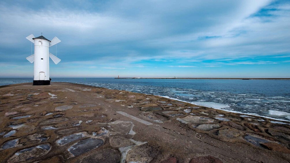 A white lighthouse on a rocky shore with the ocean and a distant red tower in the background. 5