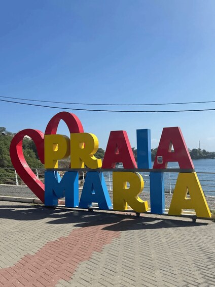 A colorful sign with the words 'Praia Maria' in large letters, set against a backdrop of a clear blue sky and a body of water. 1