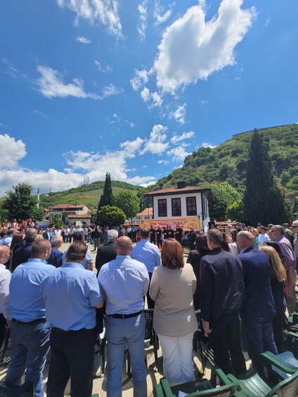 A crowd gathered outdoors, facing a building with a red-tiled roof, under a clear blue sky. 3