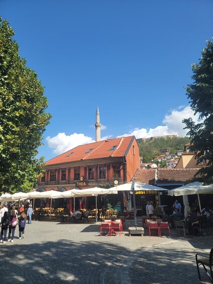 A cobblestone square with outdoor seating, a historic building labeled 'THEA HOTEL,' and a mosque with a minaret in the background. 2