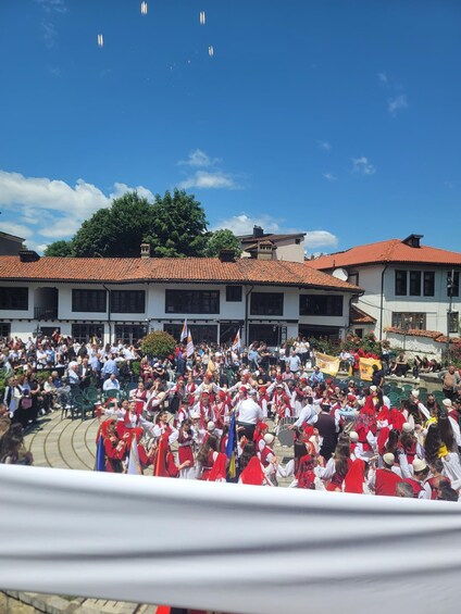 A crowd in traditional attire gathered in an outdoor square with buildings in the background. 4