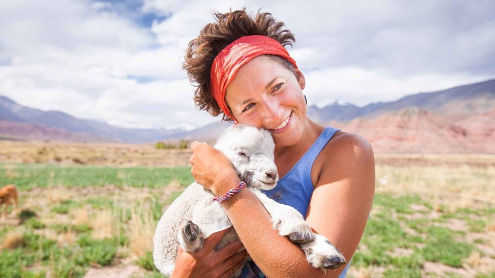 A person holding a lamb in a grassy field with mountains in the background. 5