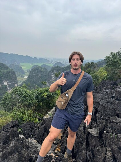 A person giving a thumbs-up gesture on a rocky outcrop with a mountainous landscape in the background. 4