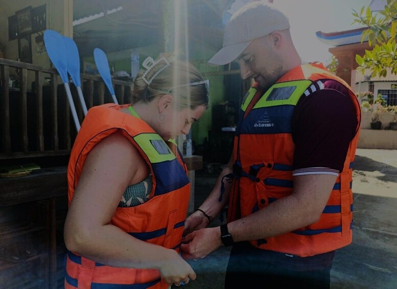 Two people in orange life vests, one wearing a cap, standing indoors with a window and a paddle in the background. 5