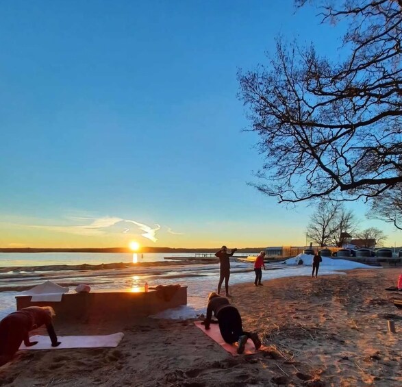 A beach scene with people exercising on mats, a sunset in the background, and a tree to the right. 2