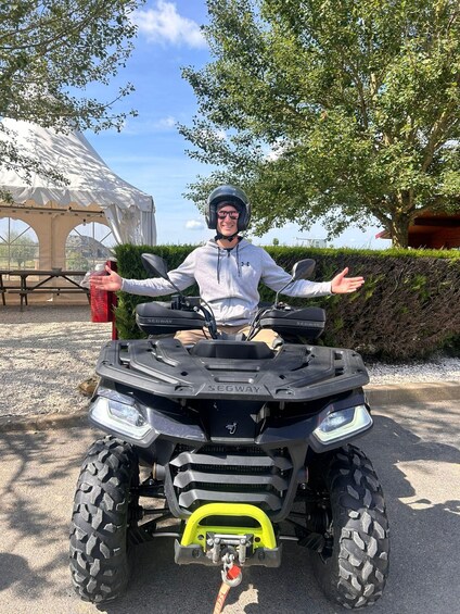 A person on a Segway ATV with a yellow front guard, standing on a paved surface with trees and a white tent in the background. 3