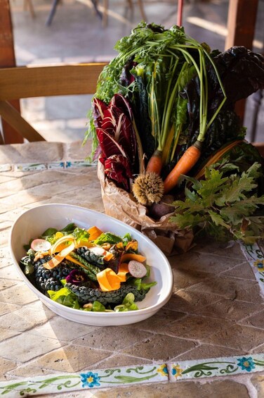 A salad bowl on a tiled table with a variety of vegetables and a basket of fresh produce in the background. 2