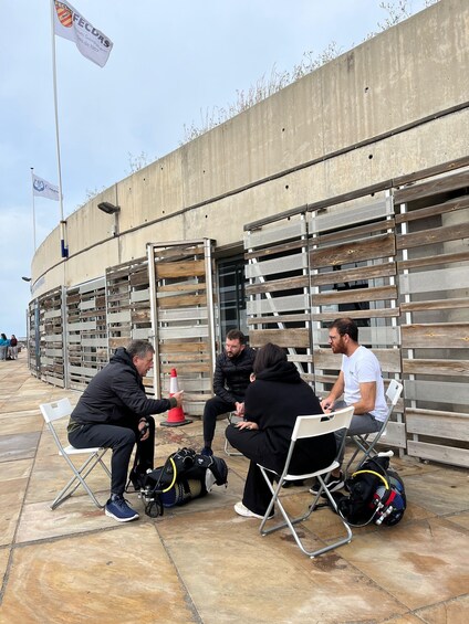 Four people seated on folding chairs outdoors, with a concrete wall and metal racks in the background. 2