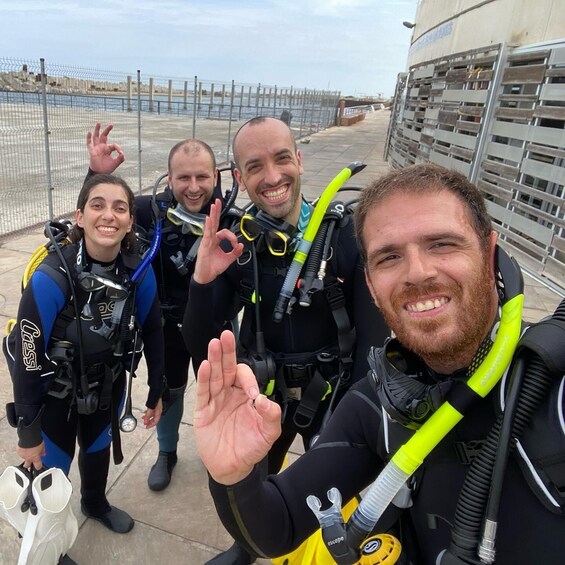 Four people in scuba diving gear with a metal fence and a building in the background. 1