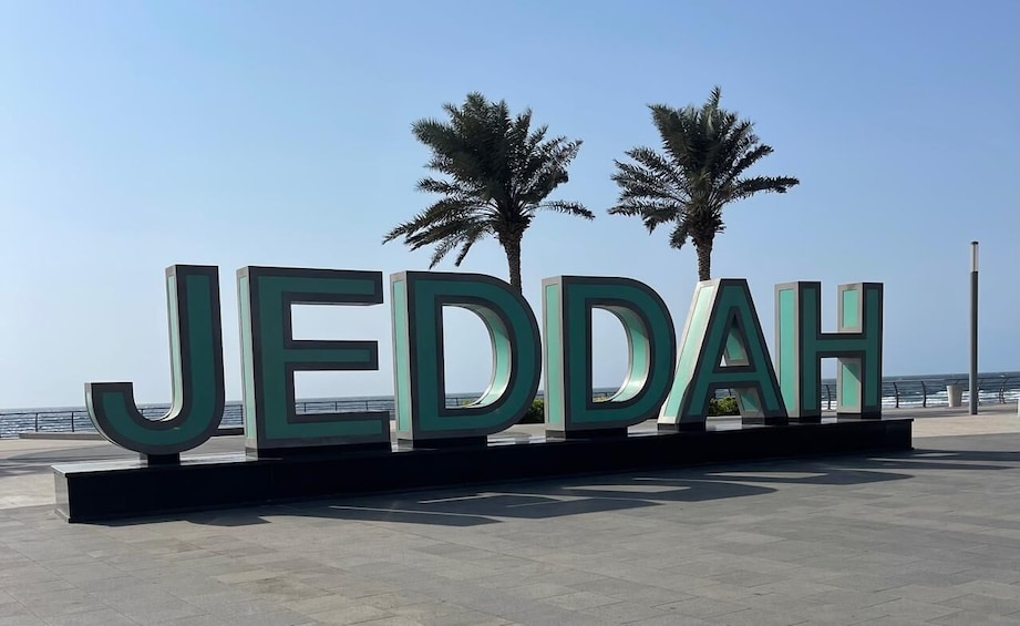 Large green letters spelling 'JEDDAH' on a platform with palm trees and a clear sky in the background. 1