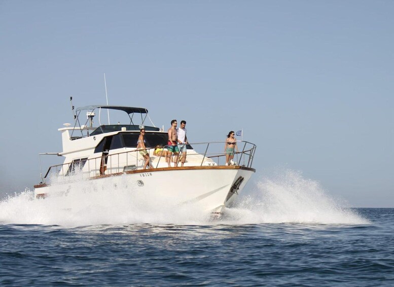 A white yacht with three people on deck, one standing and two sitting, moving through the water. 1