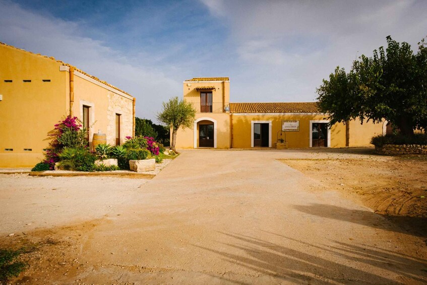 A yellow building with a small tower, surrounded by a dirt courtyard and flowering plants. 2