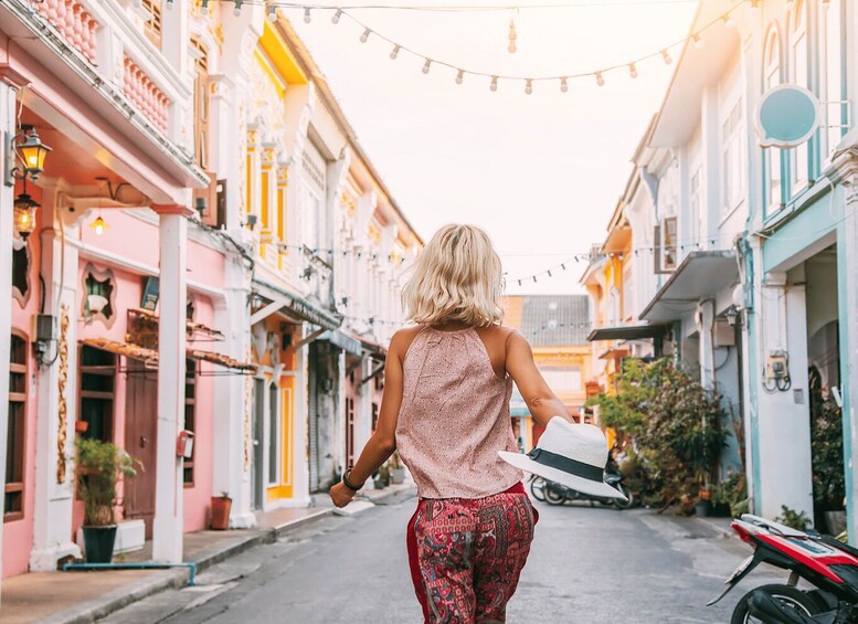 A woman walking down a narrow street lined with colorful buildings, holding a hat. 3