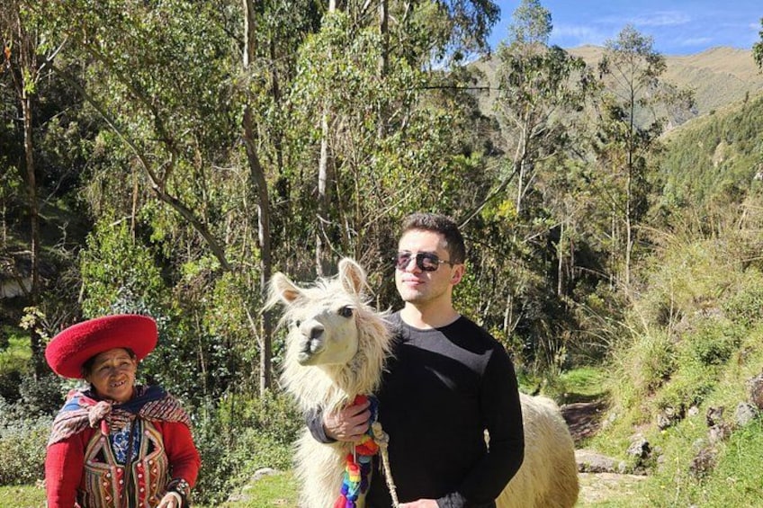 A person in a red hat and traditional clothing standing next to a white llama in a forested area with hills in the background. 1