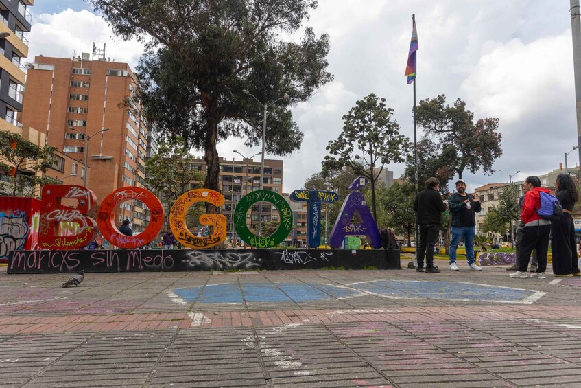 A public square with large, colorful letters spelling 'BOGOTA' on a wall, surrounded by buildings and trees. 4