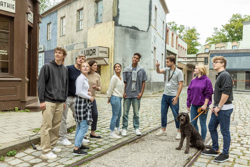 A group of people standing on a cobblestone street with a dog, in front of a building with a sign that reads 'LUXOR HOTEL'. 1
