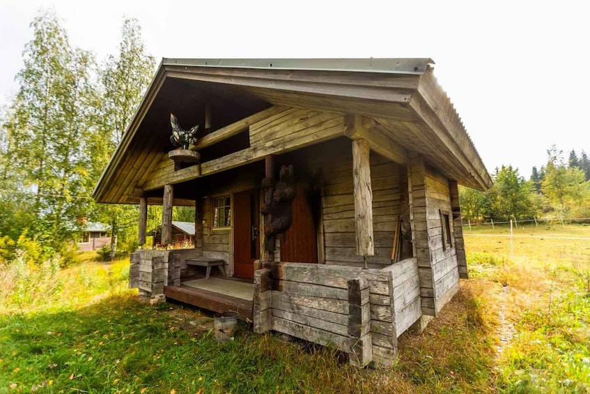 A rustic wooden cabin with a porch, surrounded by a grassy field and trees. 1